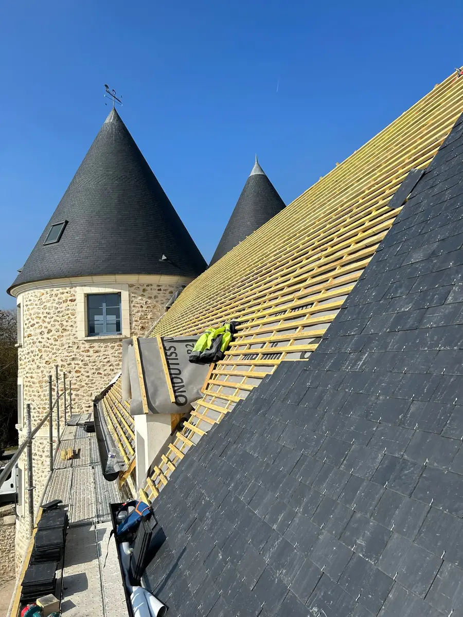 Versant de toiture en cours de couverture : à droite l'ardoise déjà posée et fixée au crochet, à gauche le liteaunage encore nu sur l'écran sous-toiture, paquets d'ardoises stockés sur l'échafaudage.