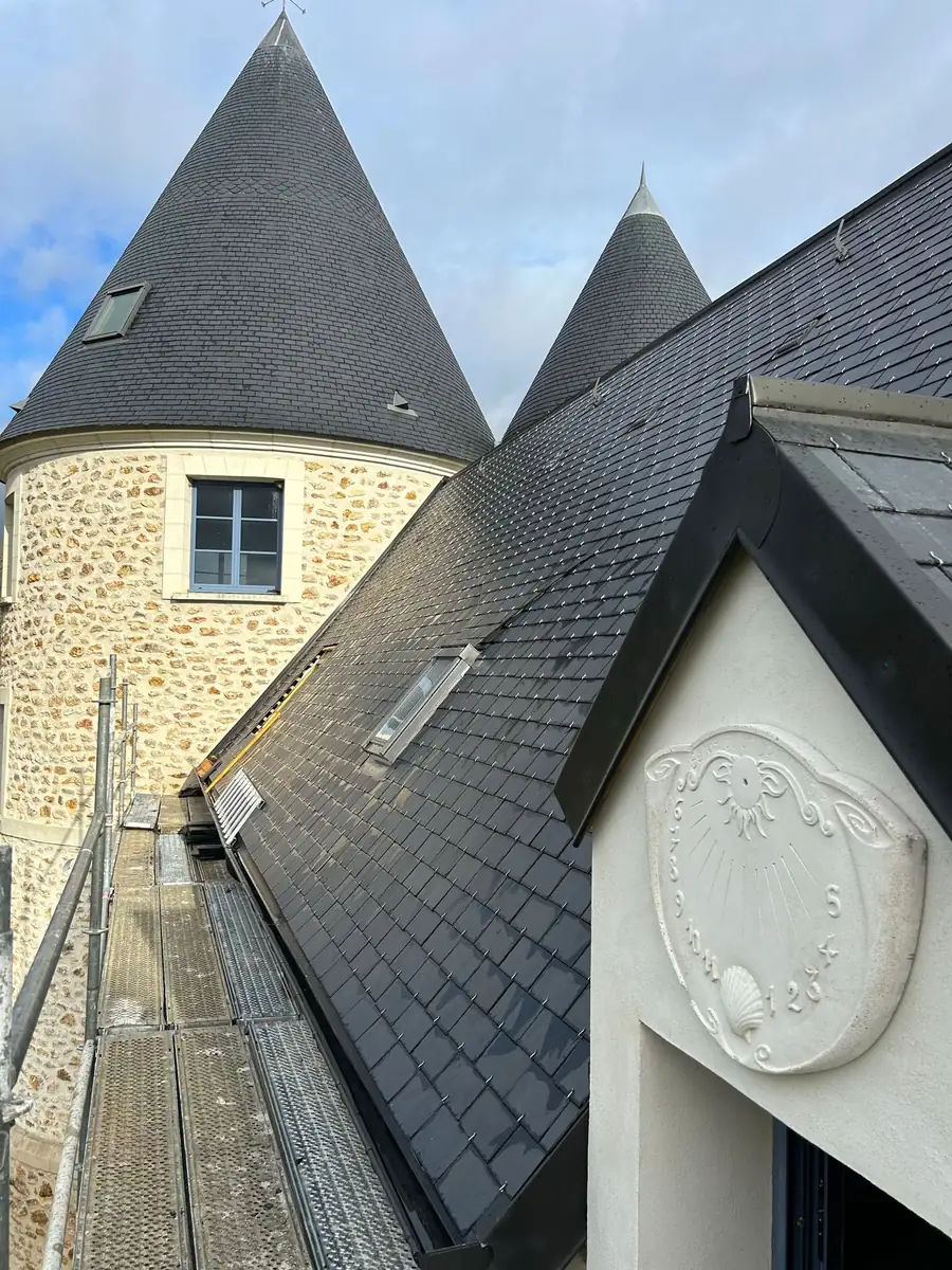 Toiture terminée en ardoise naturelle posée au crochet, arêtiers et noues habillés en zinc, fenêtres de toit intégrées et arrêts de neige alignés, avec les deux tours en poivrière et le cadran solaire de la lucarne.