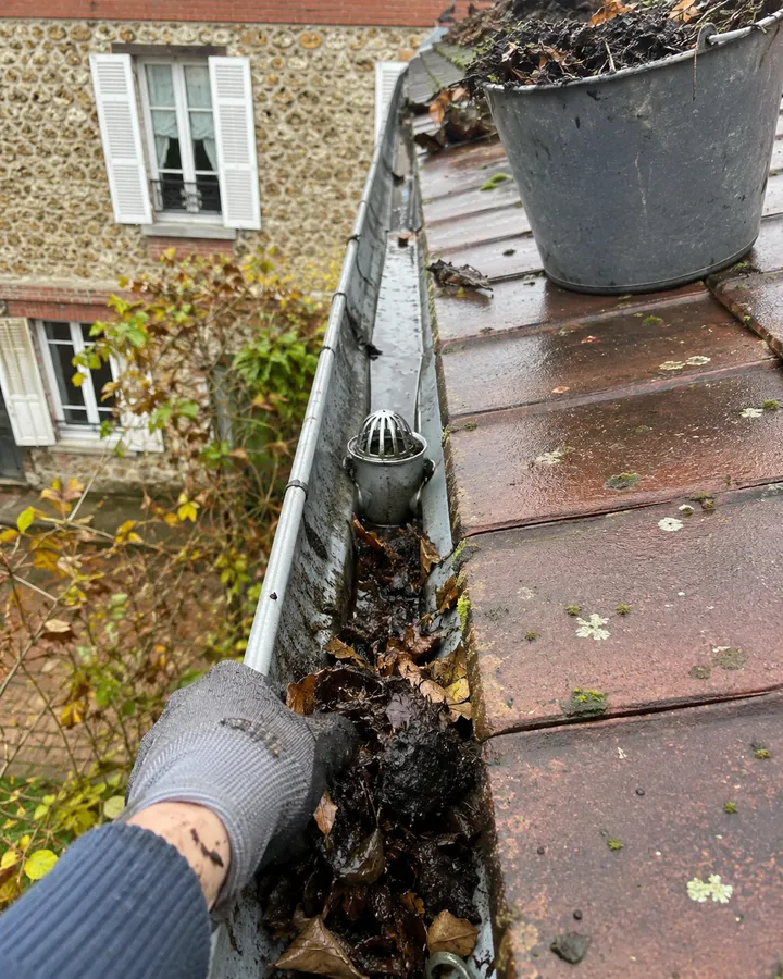 Main gantée retirant feuilles et mousse noire accumulées dans un chéneau en zinc, crapaudine visible et seau de débris sur le toit.